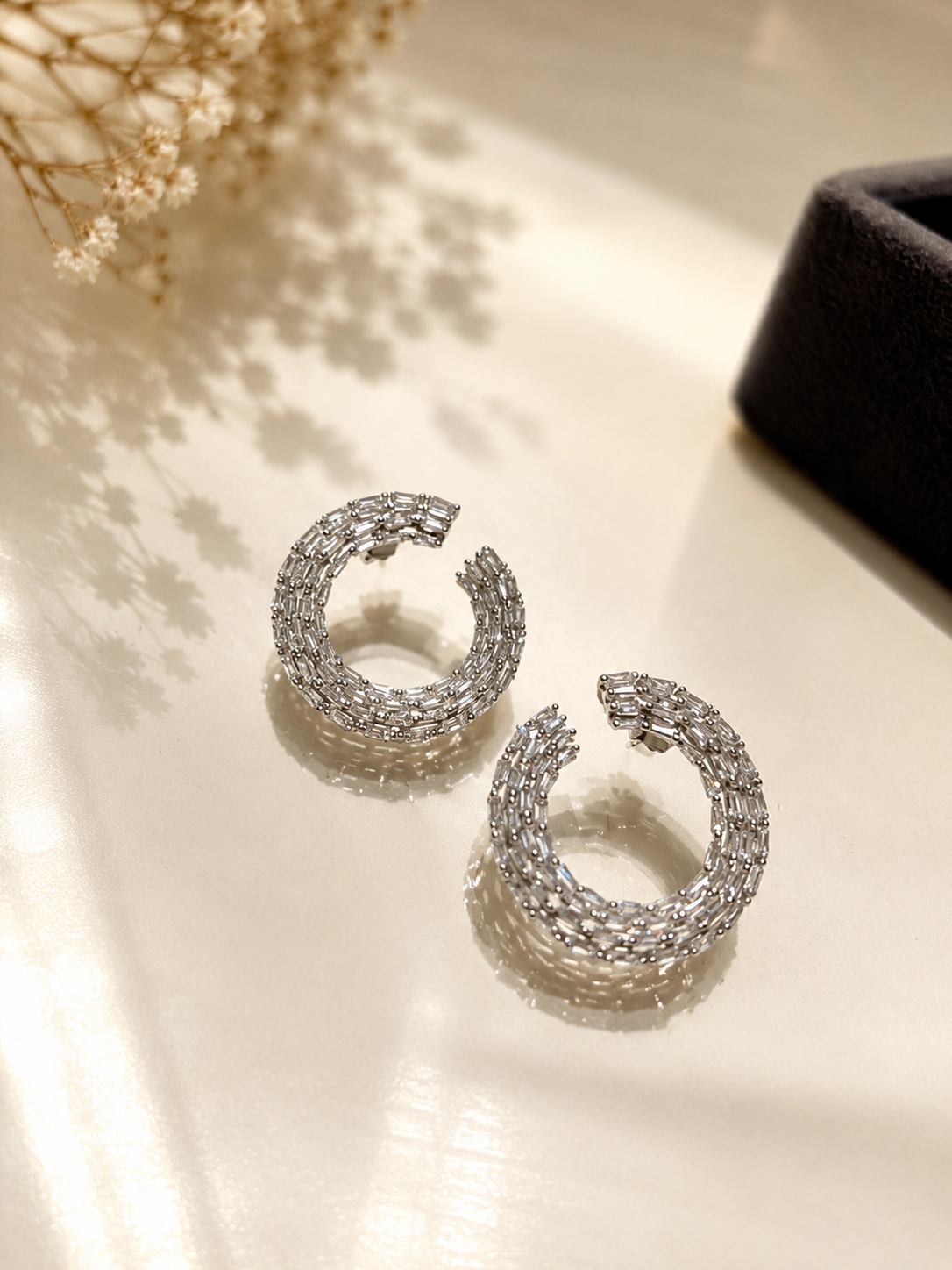 A pair of silver-toned crescent-shaped hoop earrings covered in small clear stones, displayed on a warm beige surface with soft shadows and dried flowers in the background.