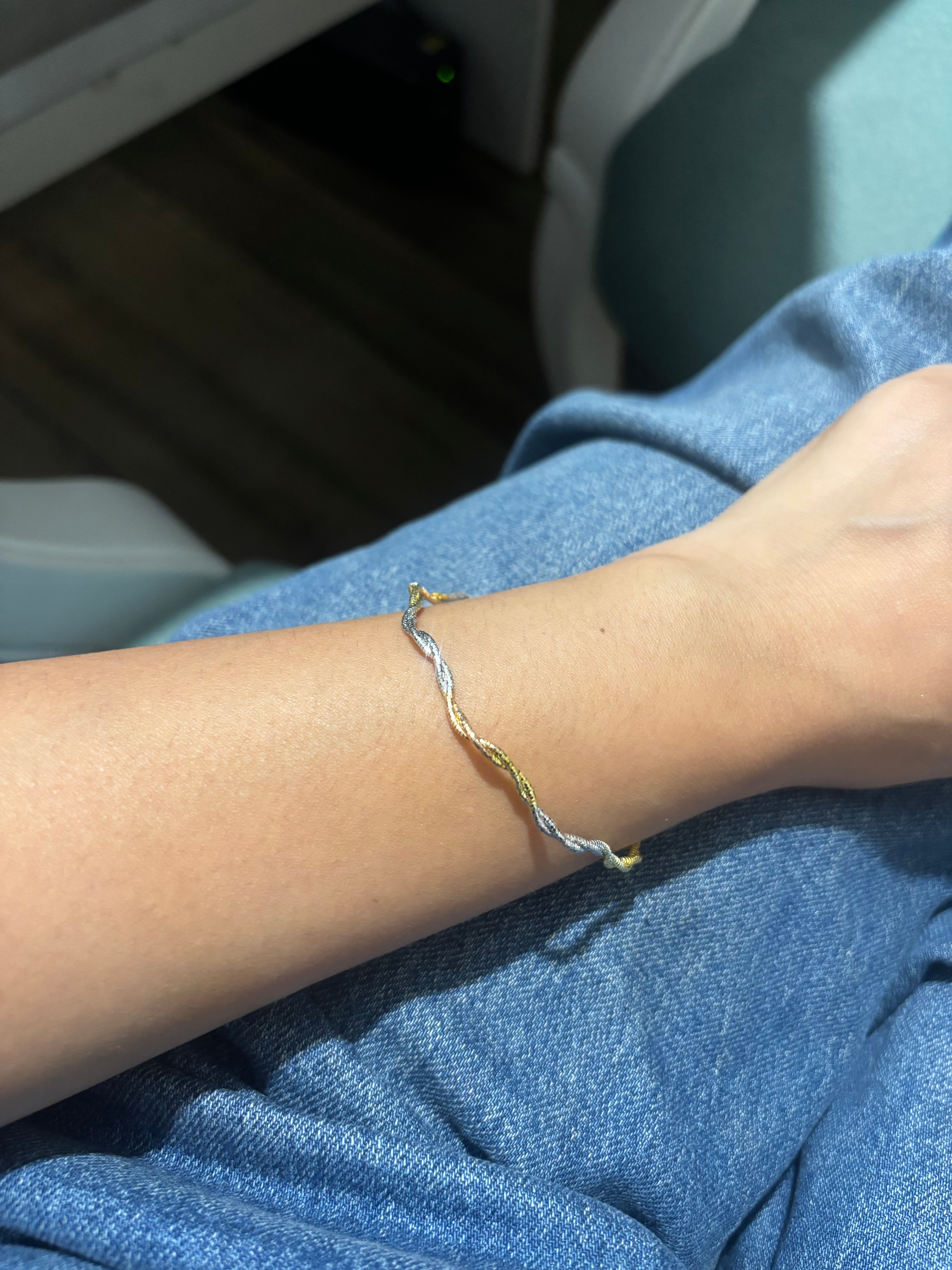 A thin twisted cord bracelet in silver, gold, and blue tones worn on a person's wrist, shown against blue denim clothing.