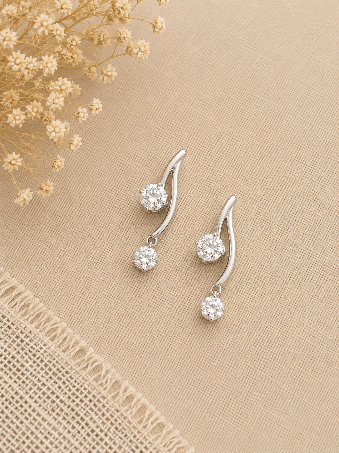 A pair of silver-toned drop earrings with curved tops and round clear gemstones, displayed on a beige textured fabric background with dried flowers in the corner.