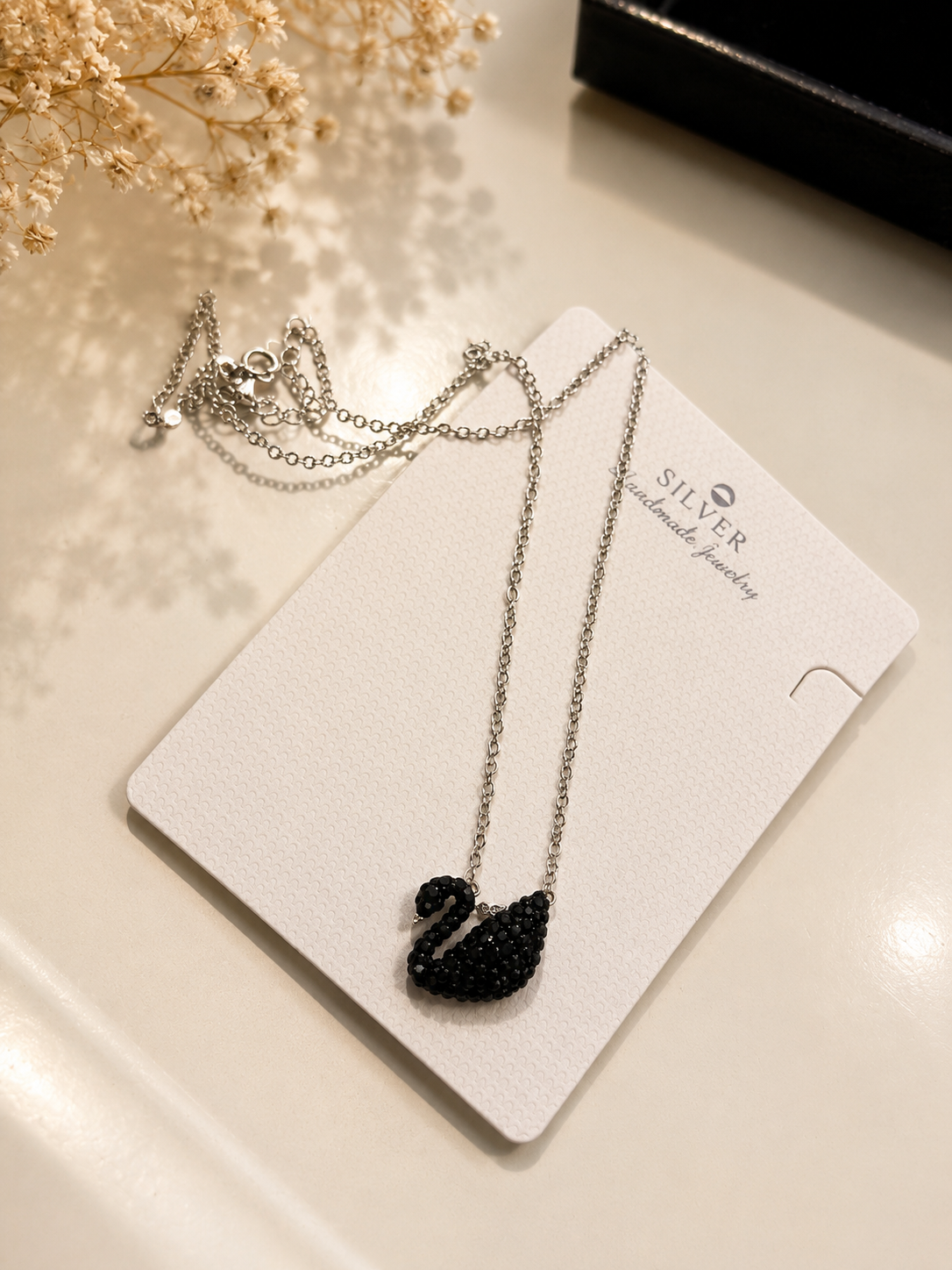 A silver-toned necklace with a black swan pendant, displayed on a white jewelry card labeled 'SILVER handmade jewelry' on a light beige surface with dried flowers in the background.