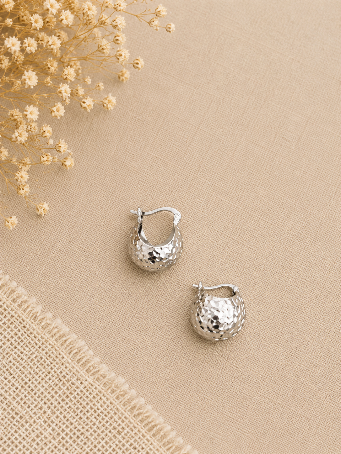 A pair of silver textured hoop earrings with a hammered finish, displayed on a beige linen fabric background with dried white flowers in the top-left corner.