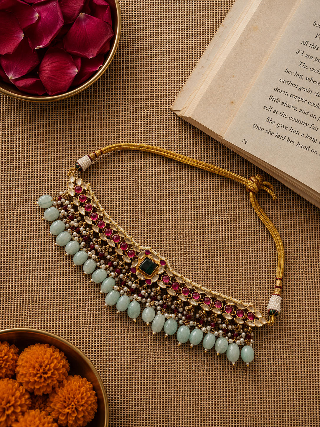 Handcrafted Kundan choker necklace set featuring ruby red stones, an emerald green center stone, and pale aqua bead drops with matching statement earrings. The gold-tone festive set is finished with pearl detailing and adjustable thread ties, styled on a soft grey velvet display.