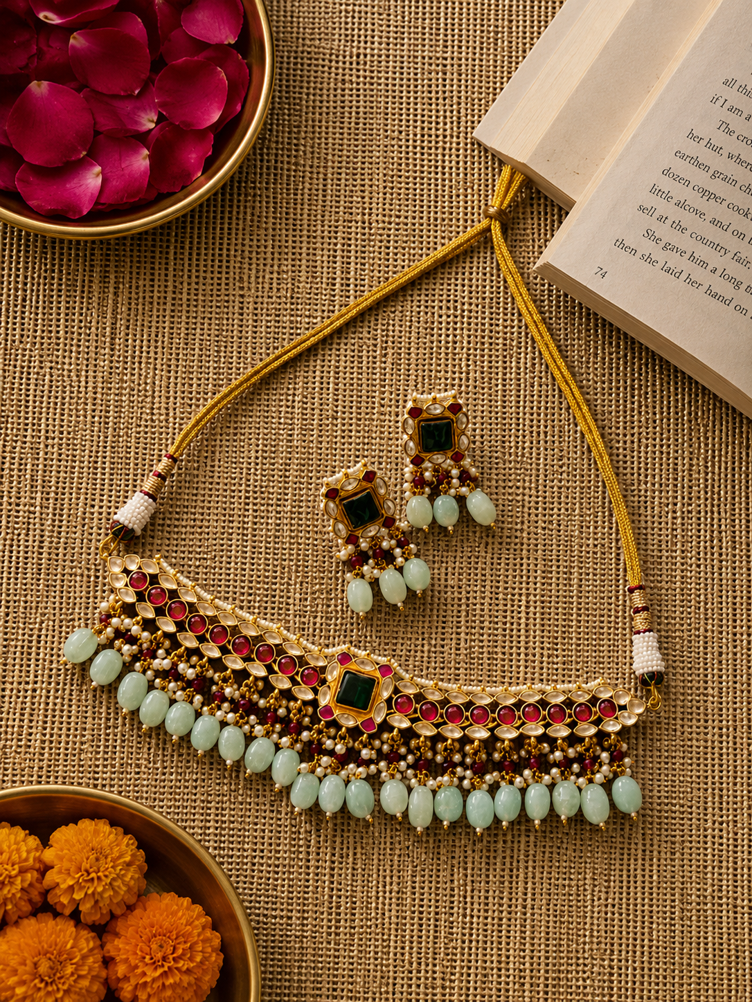 Handcrafted Kundan choker necklace set featuring ruby red stones, an emerald green center stone, and pale aqua bead drops with matching statement earrings. The gold-tone festive set is finished with pearl detailing and adjustable thread ties, styled on a soft grey velvet display.
