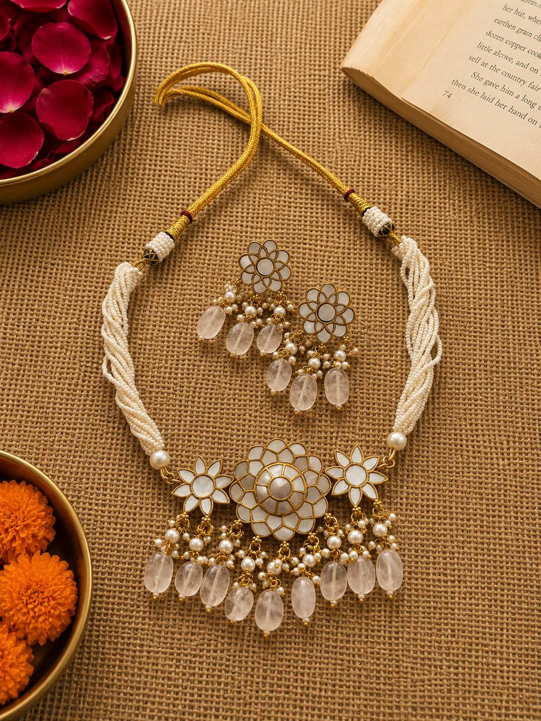 A traditional Indian jewelry set featuring a statement necklace and matching earrings. The necklace has a cream-colored braided cord, white floral enamel details, and dangling clear teardrop crystals with small pearl-like beads.
