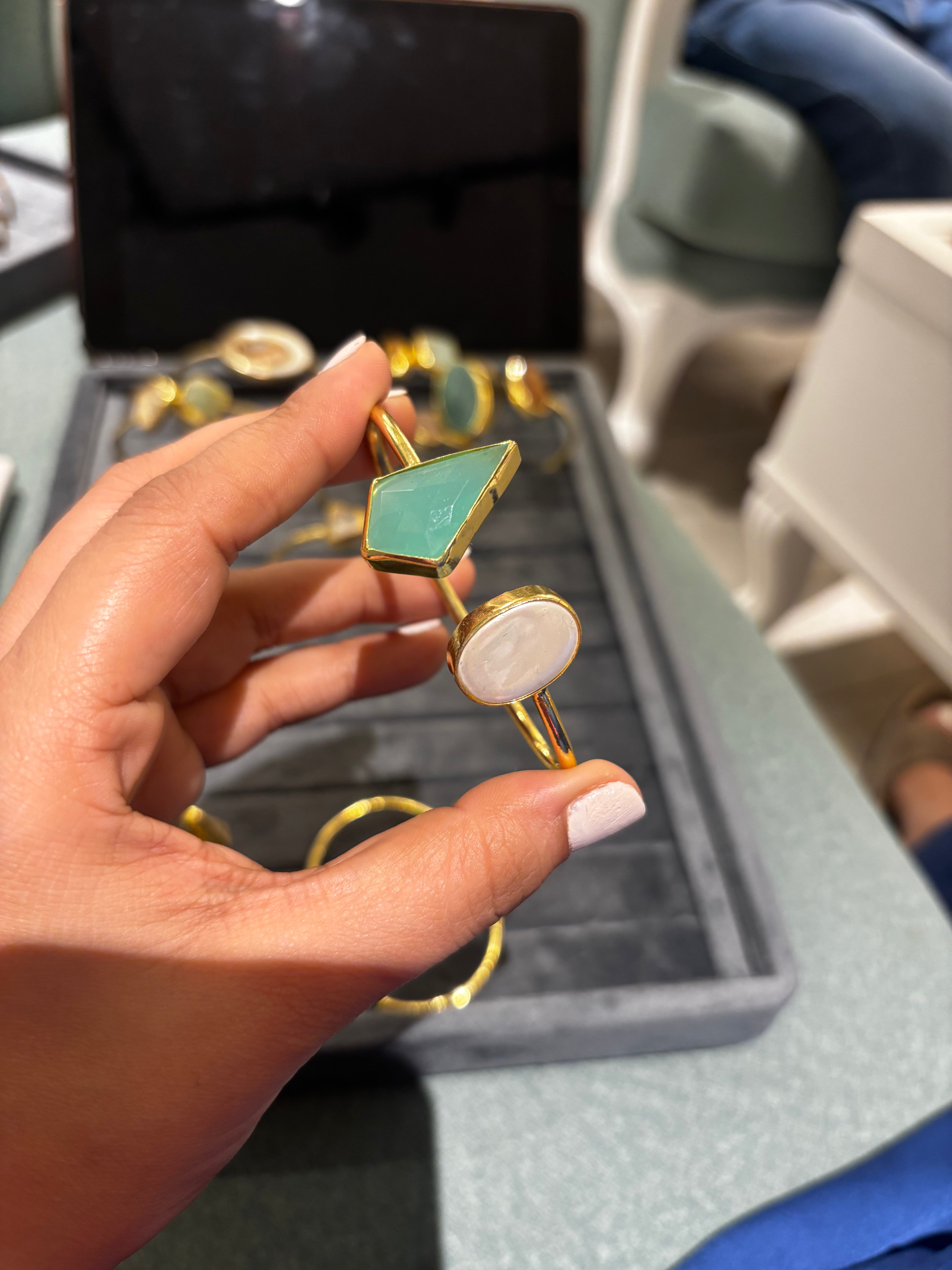 A hand holding a gold ring with a green geometric stone and a white oval stone, with a jewelry display box in the background.