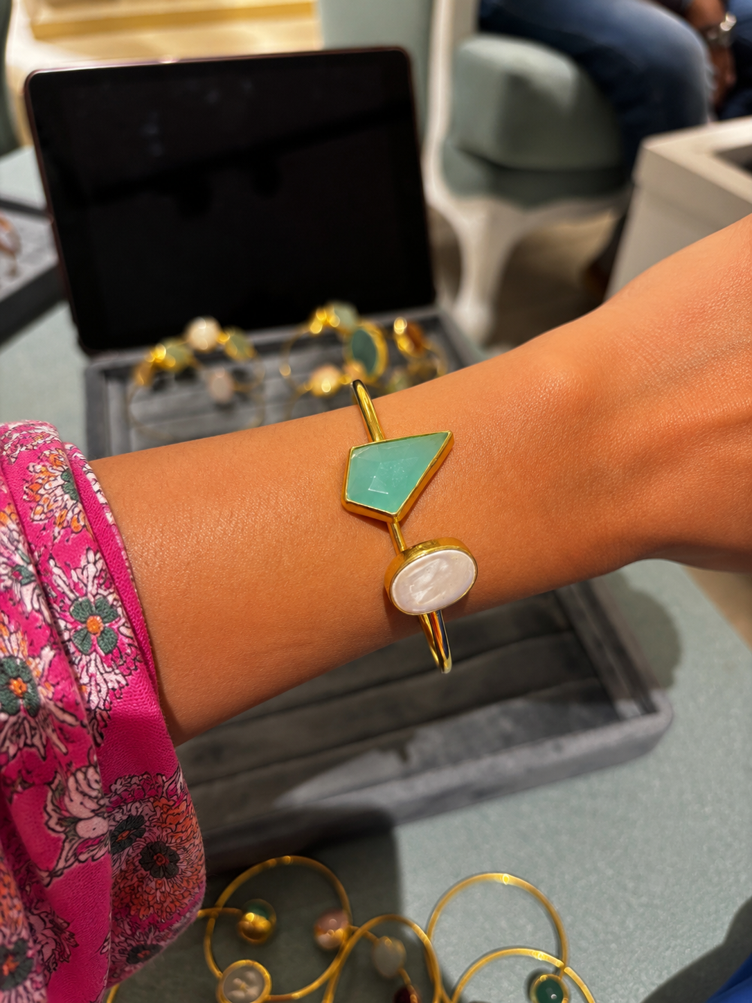 A gold bangle bracelet with a mint green geometric triangle and a white oval stone, shown worn on a person's wrist.