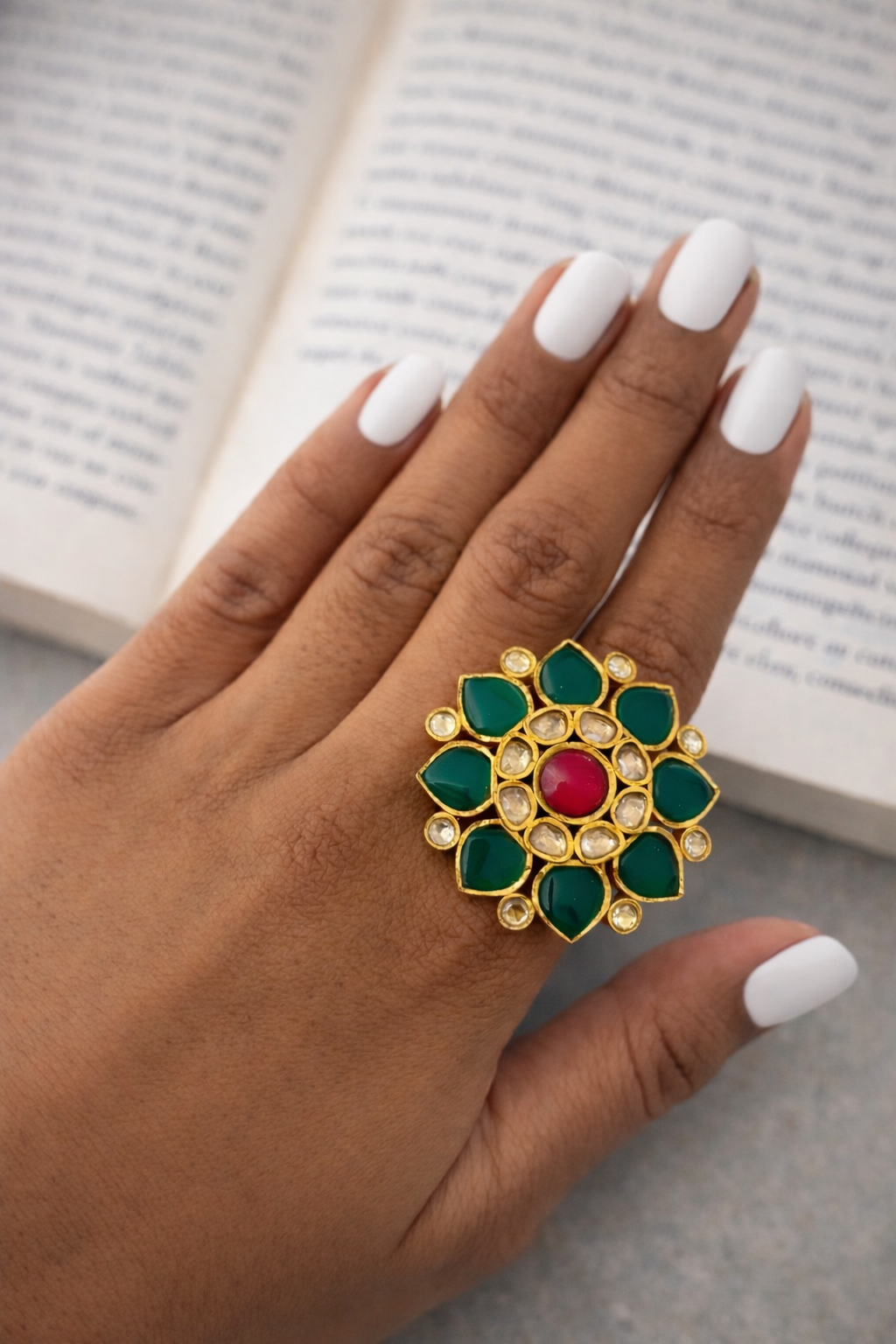 Hand wearing a decorative ring with green and red stones on a blurred book background