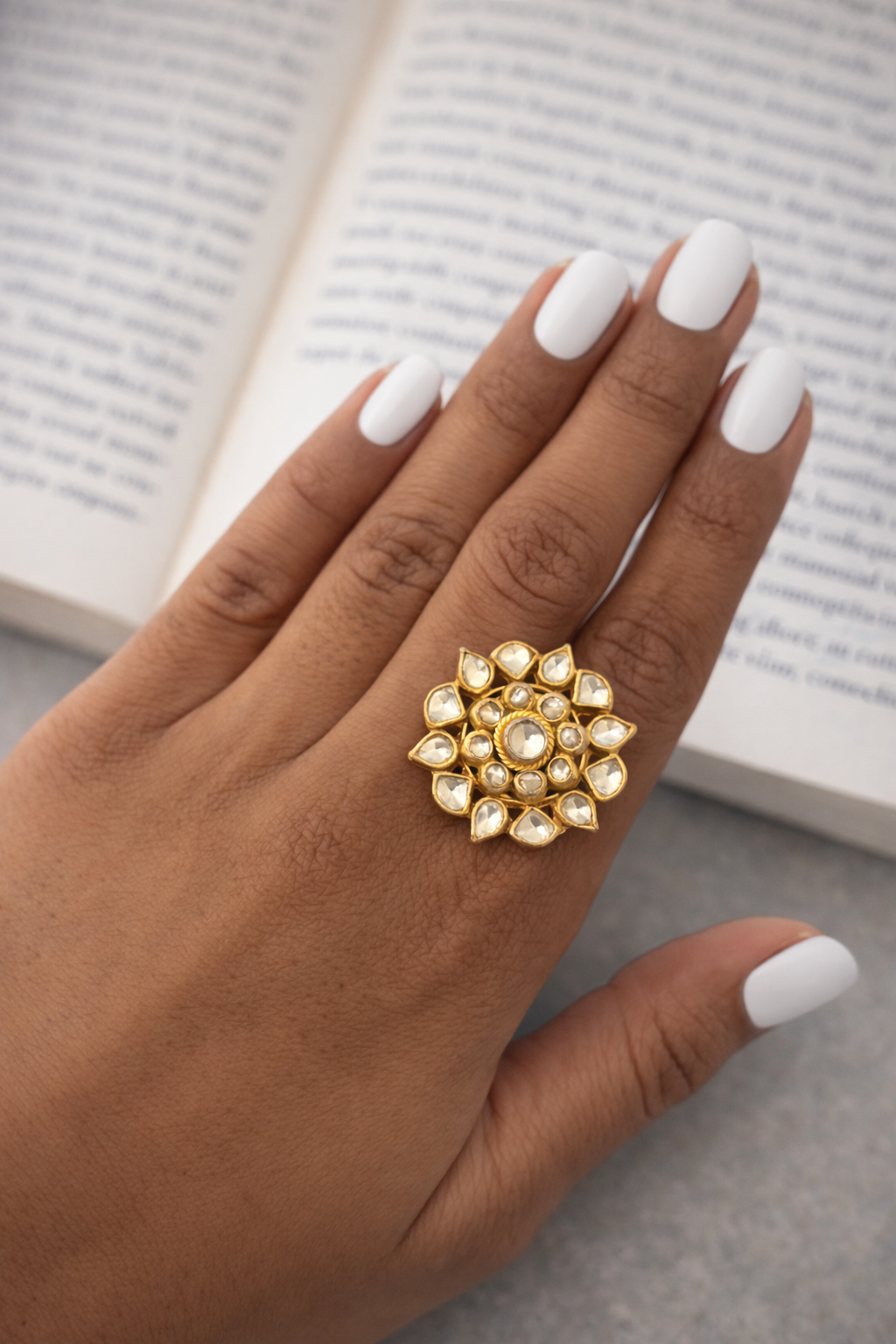 Gold ring with intricate design on a hand with white nail polish, against a blurred book background.