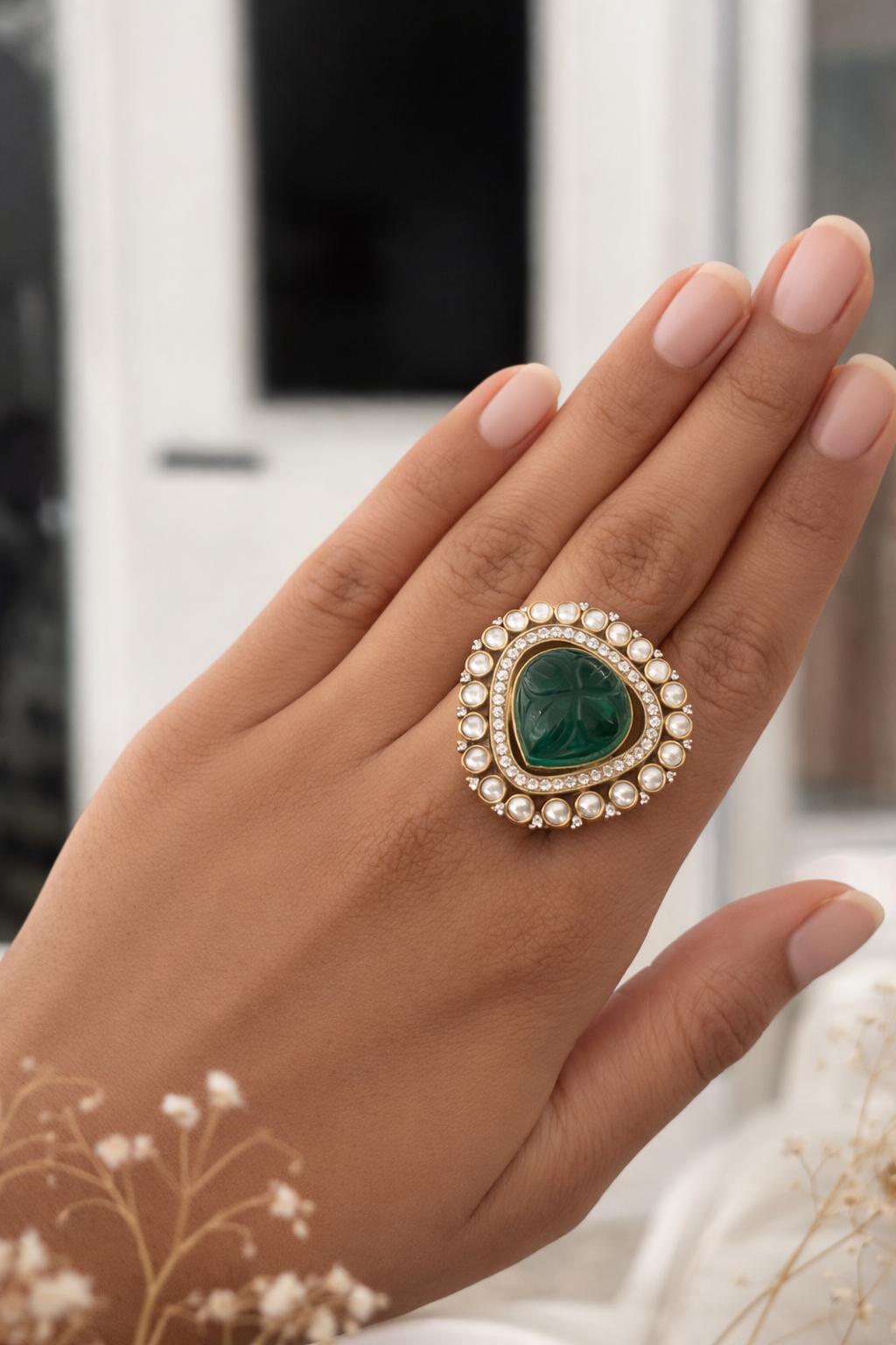 Hand wearing a ring with a green gemstone and pearl details against a blurred indoor background
