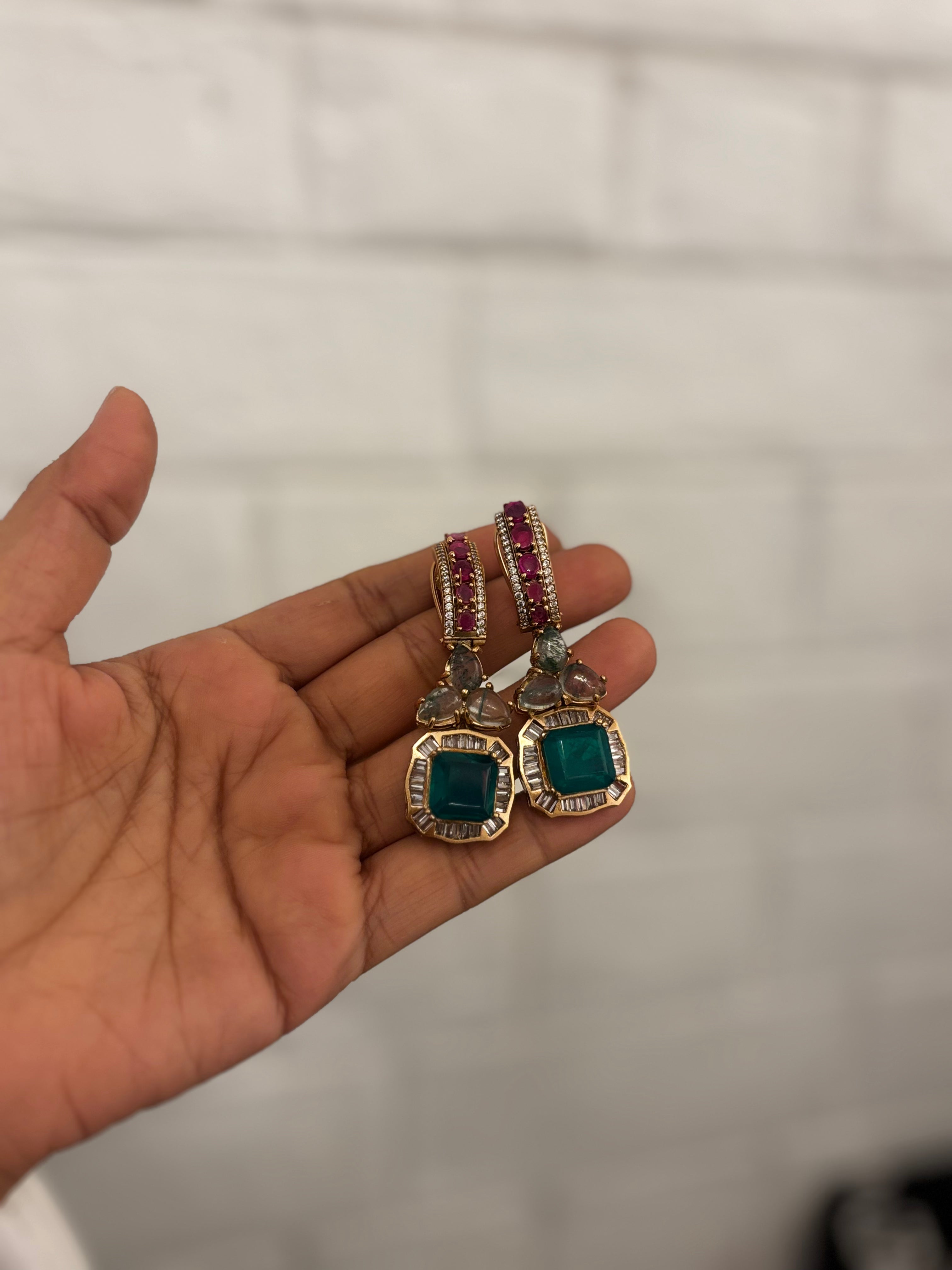 A pair of gold-plated earrings with square green gemstones and pink gemstone accents, held in a person's hand against a white brick wall background.