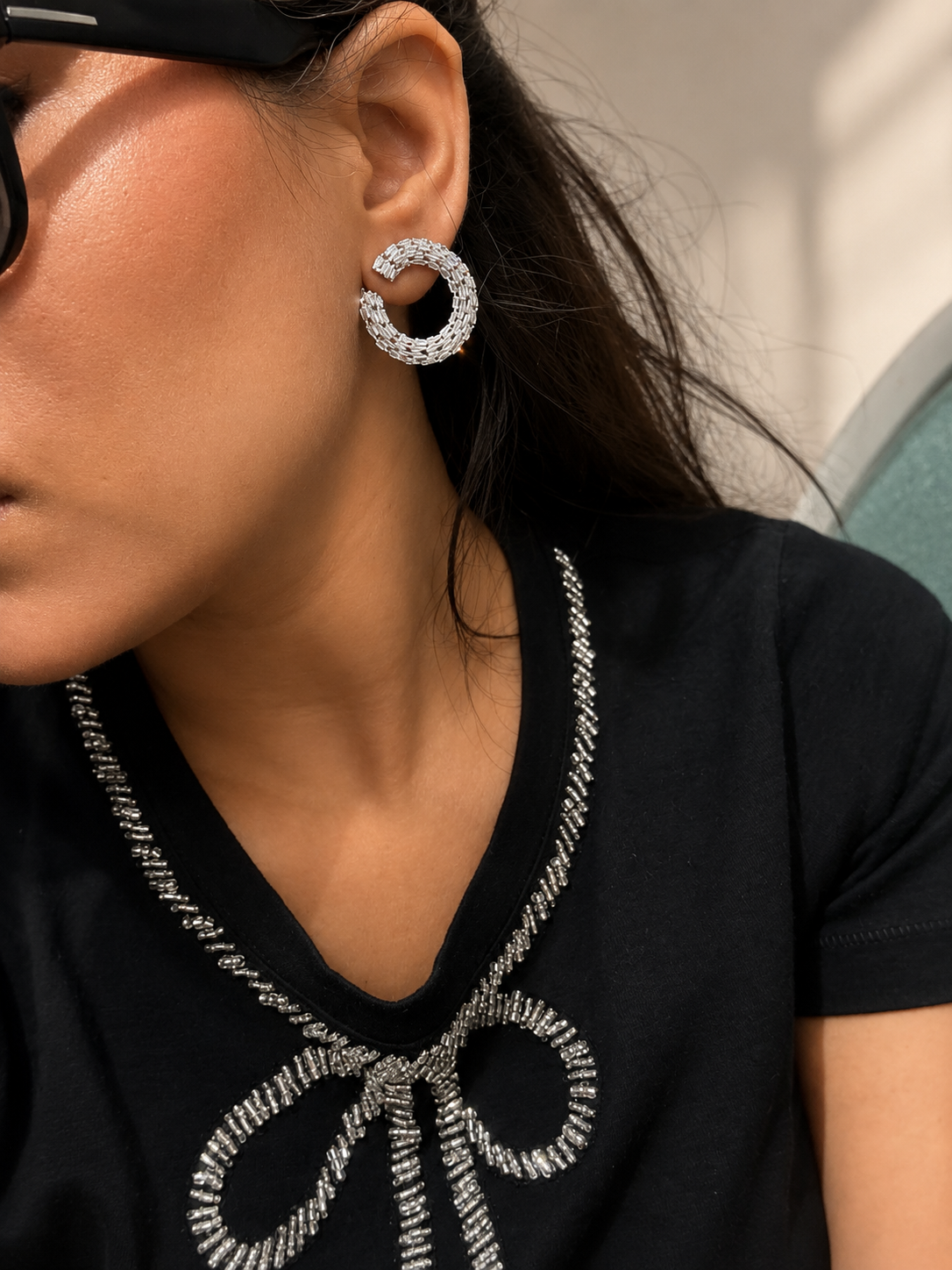 A close-up lifestyle shot of a woman wearing a sparkling silver C-shaped hoop earring with pavé stones, paired with a black t-shirt featuring silver rhinestone trim.