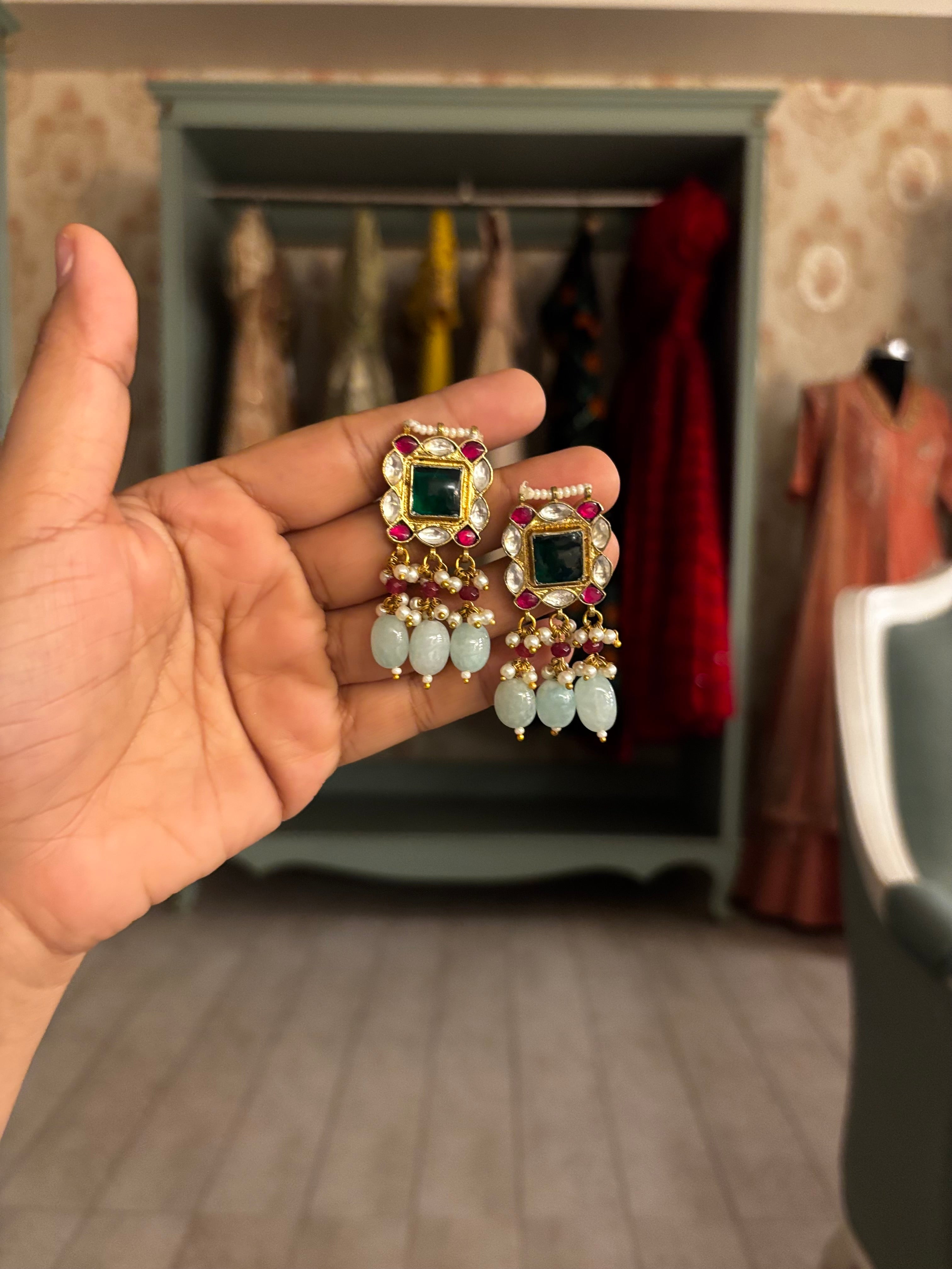 Handcrafted Kundan choker necklace set featuring ruby red stones, an emerald green center stone, and pale aqua bead drops with matching statement earrings. The gold-tone festive set is finished with pearl detailing and adjustable thread ties, styled on a soft grey velvet display.