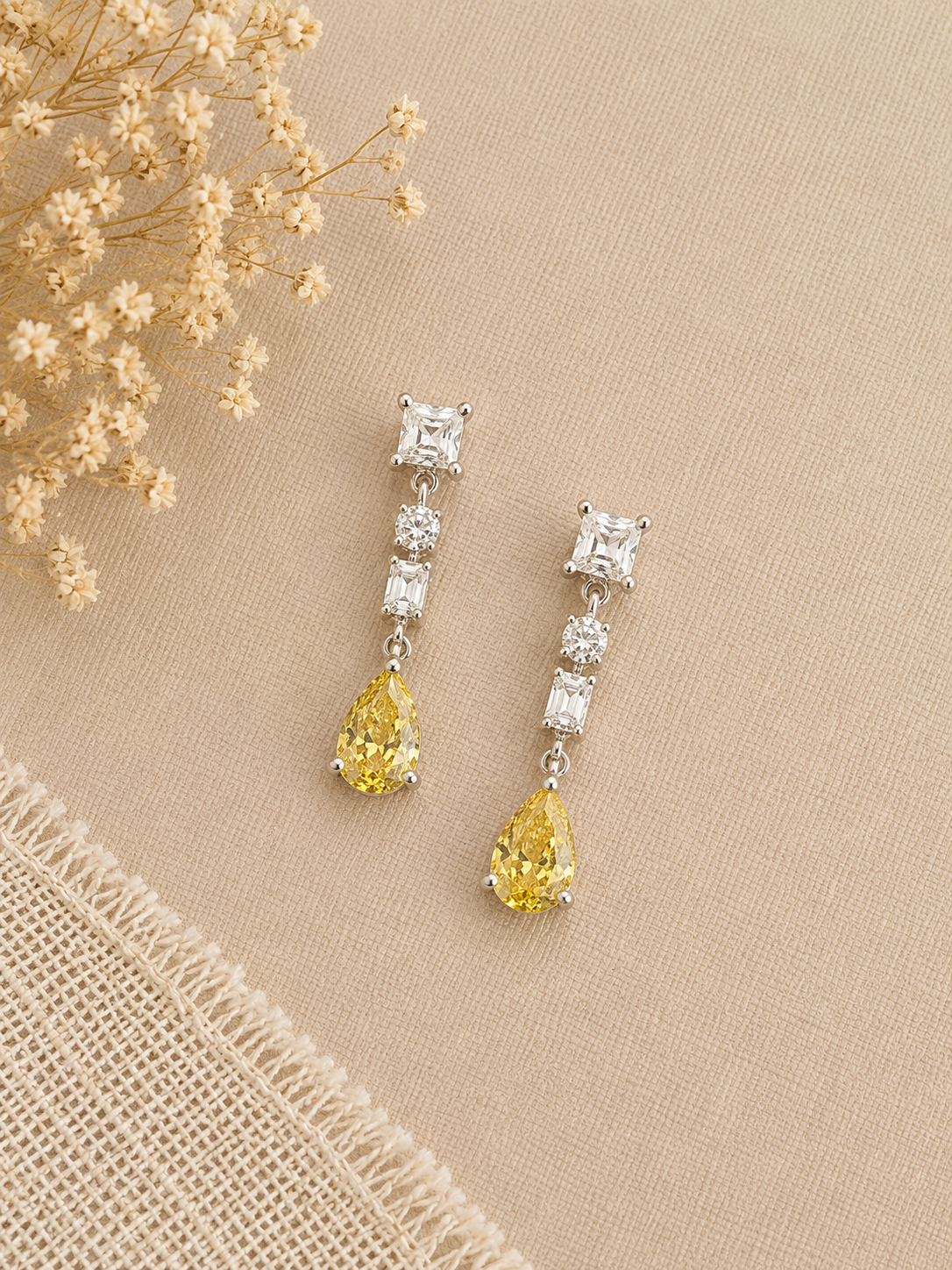 A pair of silver drop earrings with clear rectangular and round stones, each ending in a yellow teardrop gemstone, displayed on a beige textured fabric background with dried flowers.