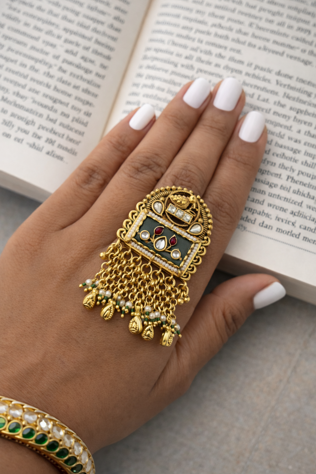 Gold ring with green gemstone and red stones on a hand with white nail polish, against an open book background.