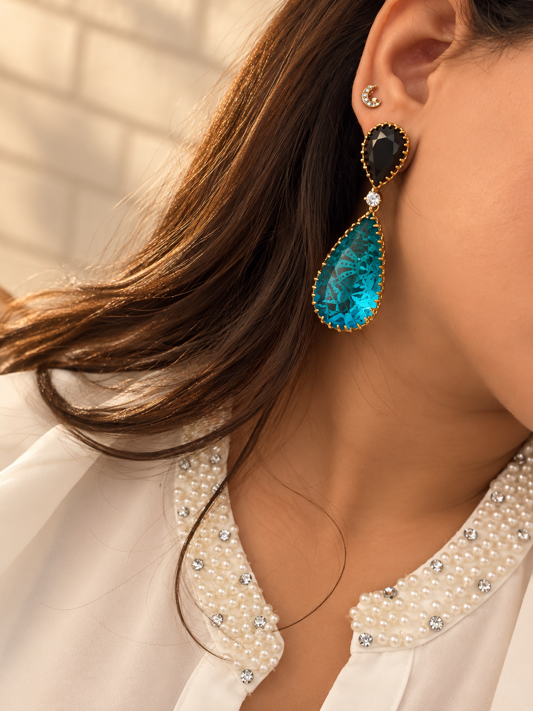 A pair of gold drop earrings with large teardrop-shaped bright blue gemstones and smaller black teardrop stones at the top, displayed on a sandy surface with dried flowers.