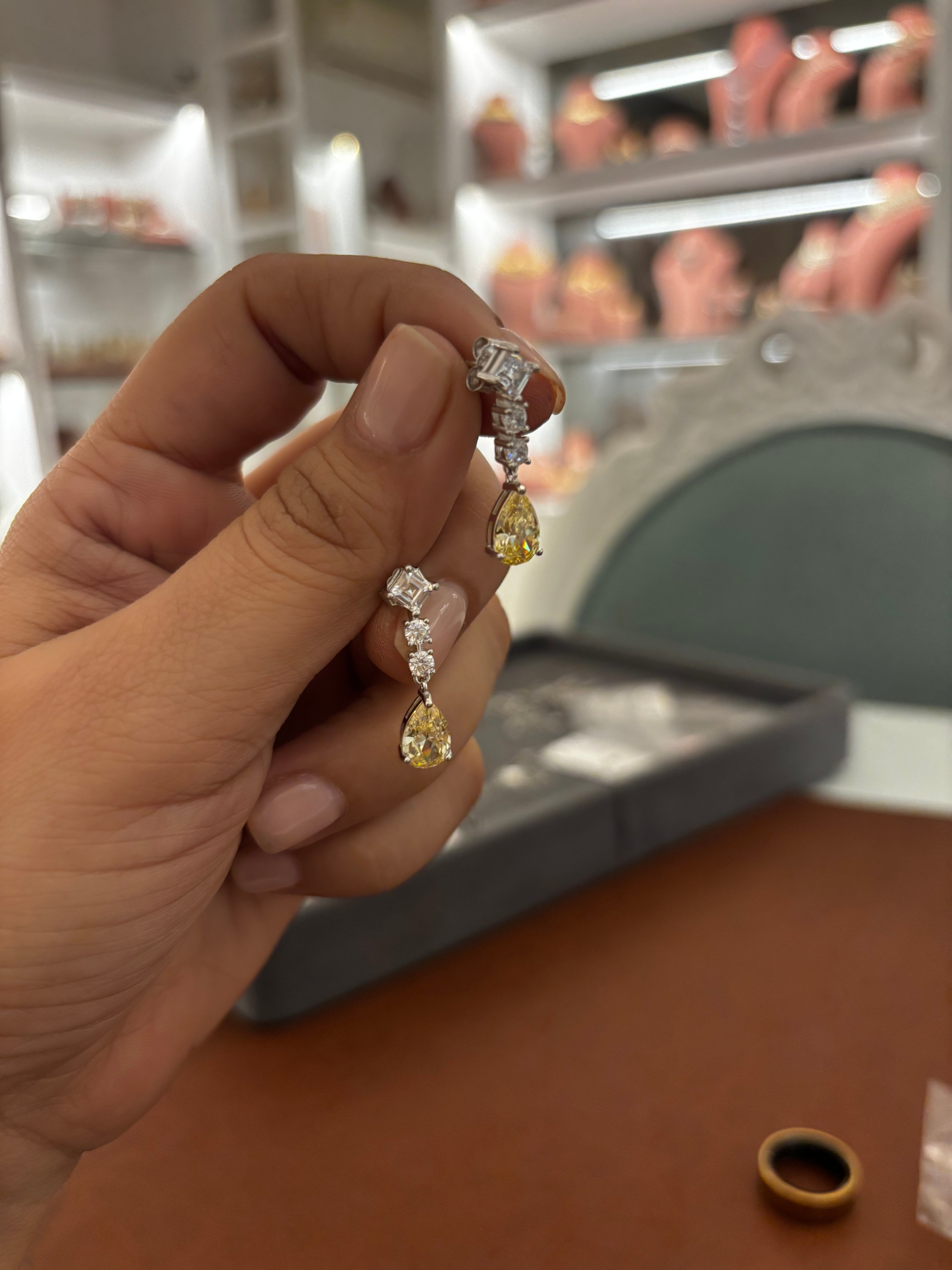 A hand holding a pair of yellow diamond drop earrings with silver settings, shown in a jewelry store setting.