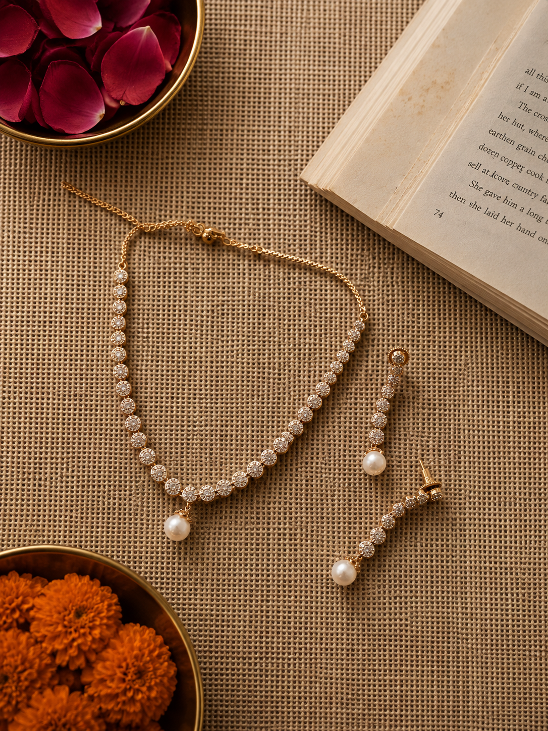 Gold-finish crystal tennis necklace with a pearl drop pendant and matching pearl drop earrings displayed on a textured beige background with floral styling.