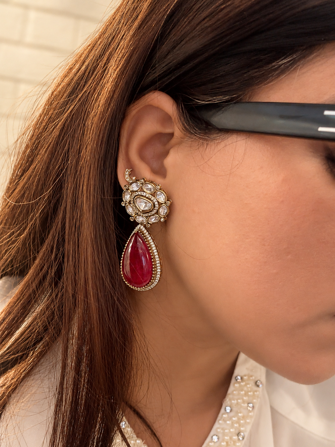 A pair of ornate gold-toned drop earrings with clear pavé-set stones at the top and large teardrop-shaped deep red stones below, displayed on a sandy beige surface with small dried flowers.