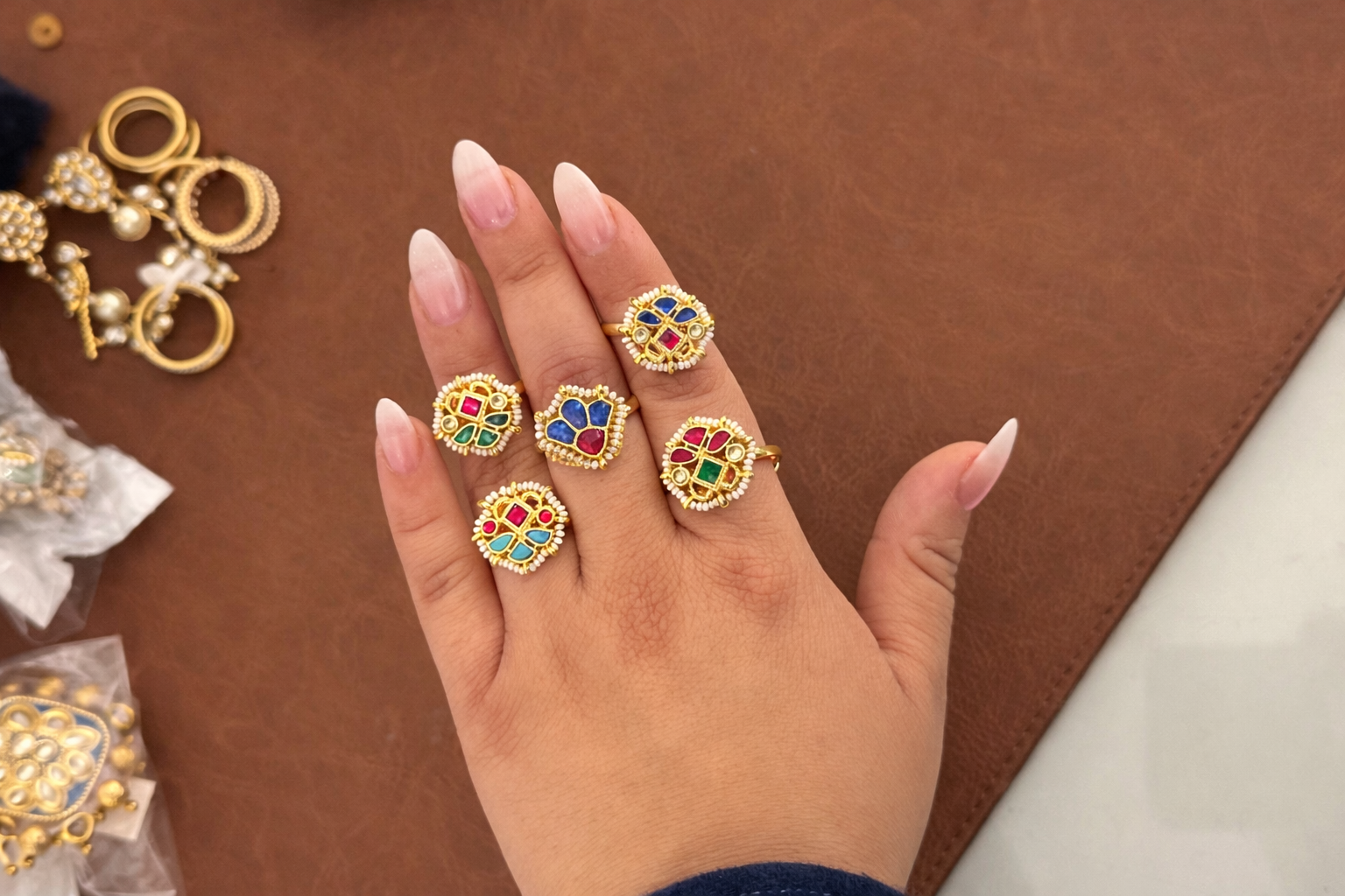Hand wearing multiple gold rings with colorful gemstones on a brown surface.