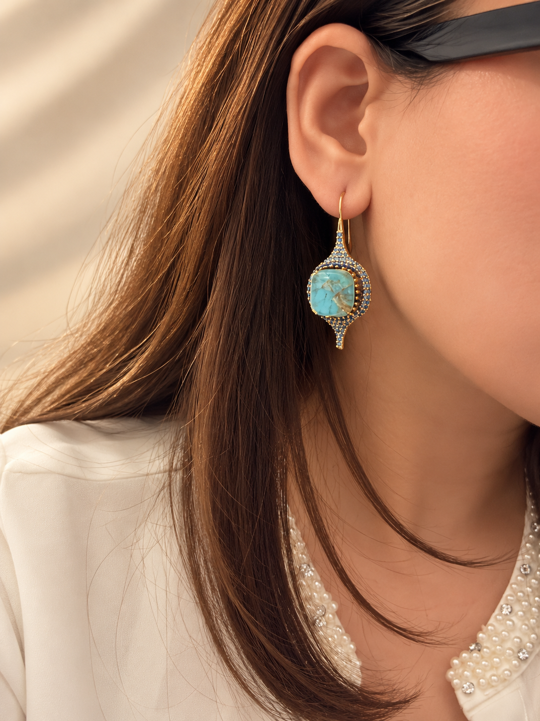 A close-up lifestyle shot of a woman wearing a gold teardrop-shaped drop earring with a light blue cabochon stone and small clear crystals.