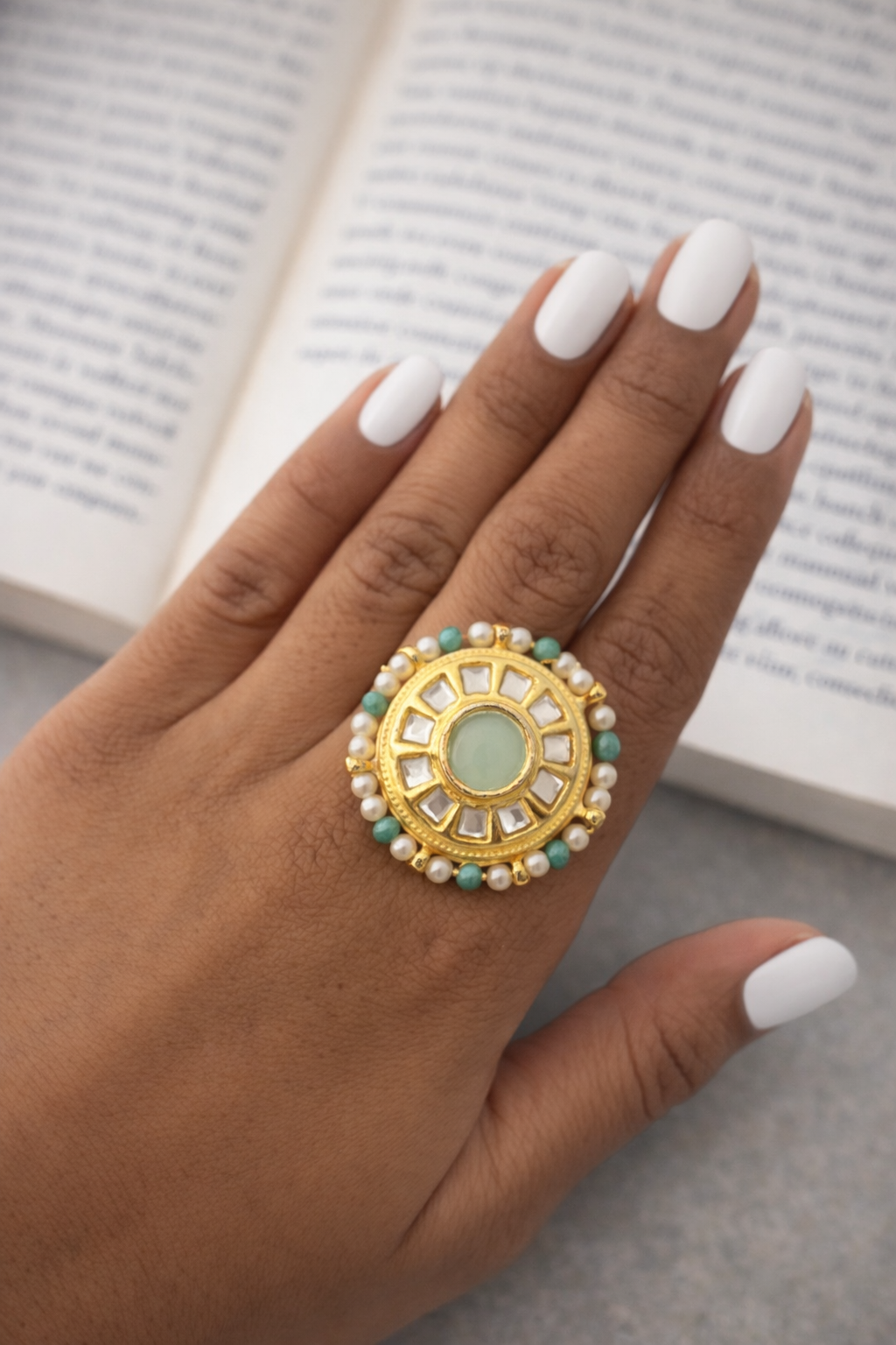 Hand wearing a decorative ring with pearls and gemstones against an open book background