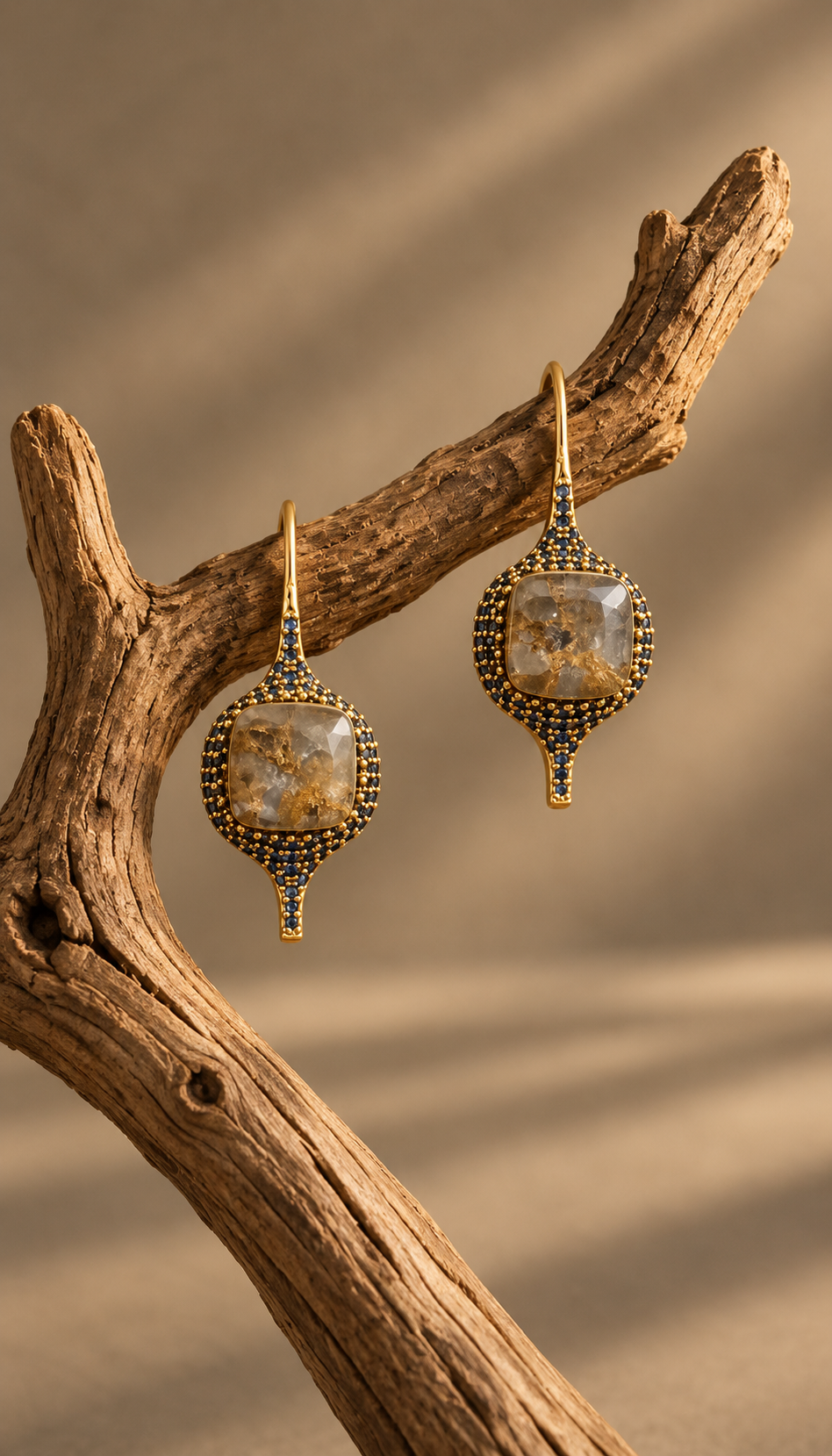 A pair of gold-toned dangle earrings with oval clear stones and dark blue beaded detailing, displayed on a piece of driftwood against a warm beige background.
