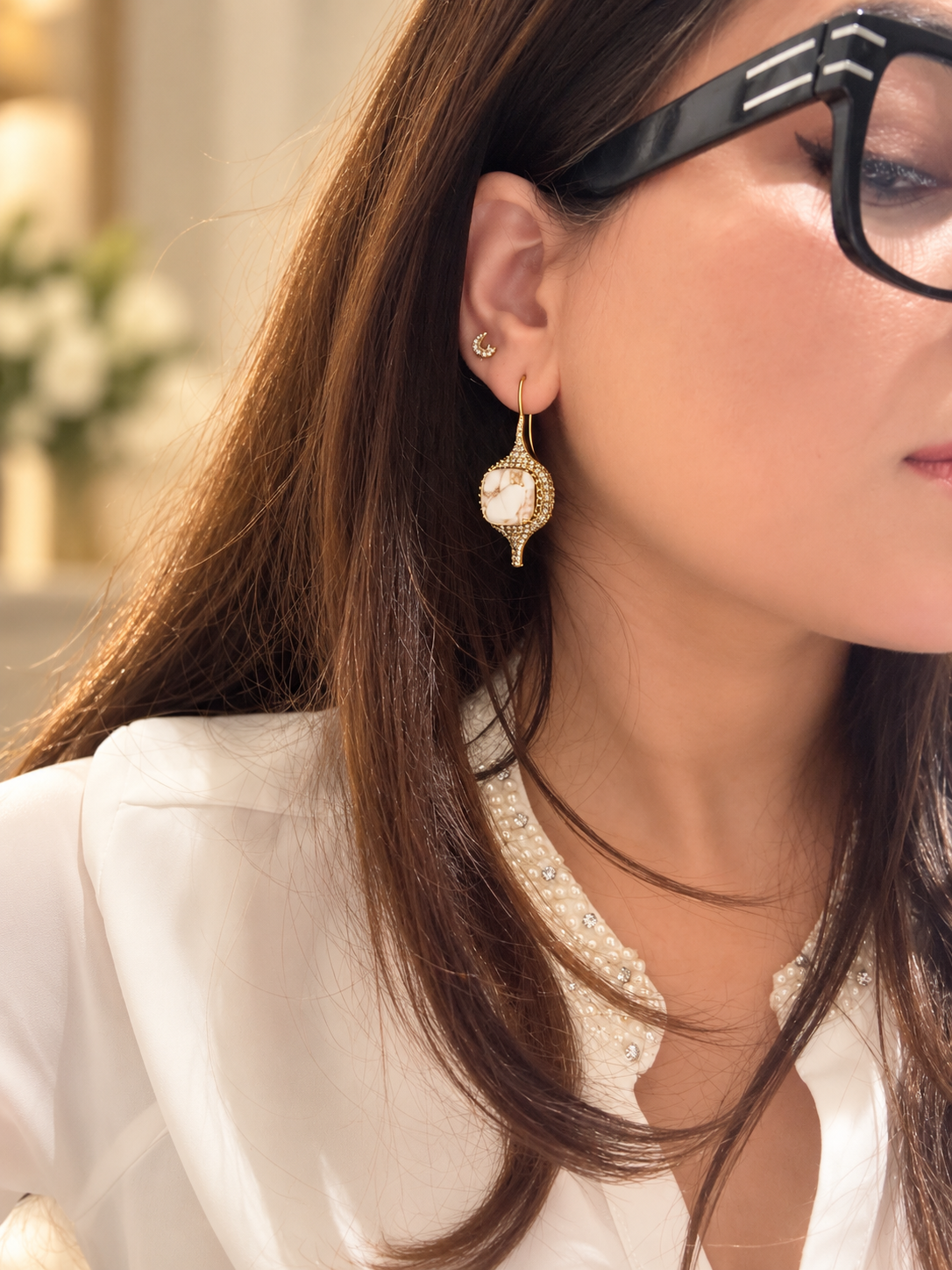 A close-up lifestyle shot of a woman wearing a gold-toned teardrop-shaped dangle earring with a white pearl center, styled with black-framed glasses and a white blouse.