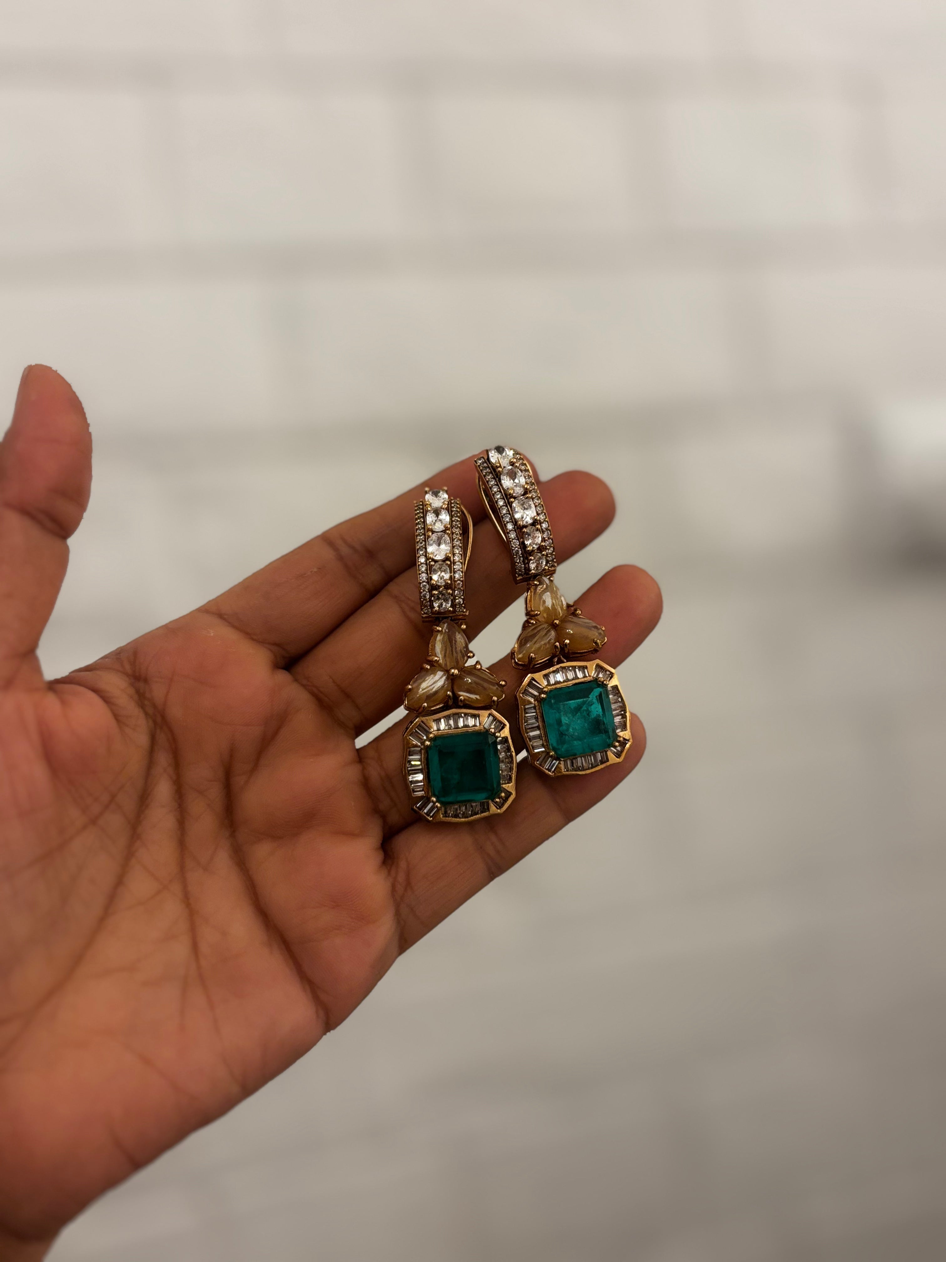 A pair of gold-plated drop earrings with square green emerald-cut stones and clear pavé-set stones, held in a person's hand against a blurred light gray background.