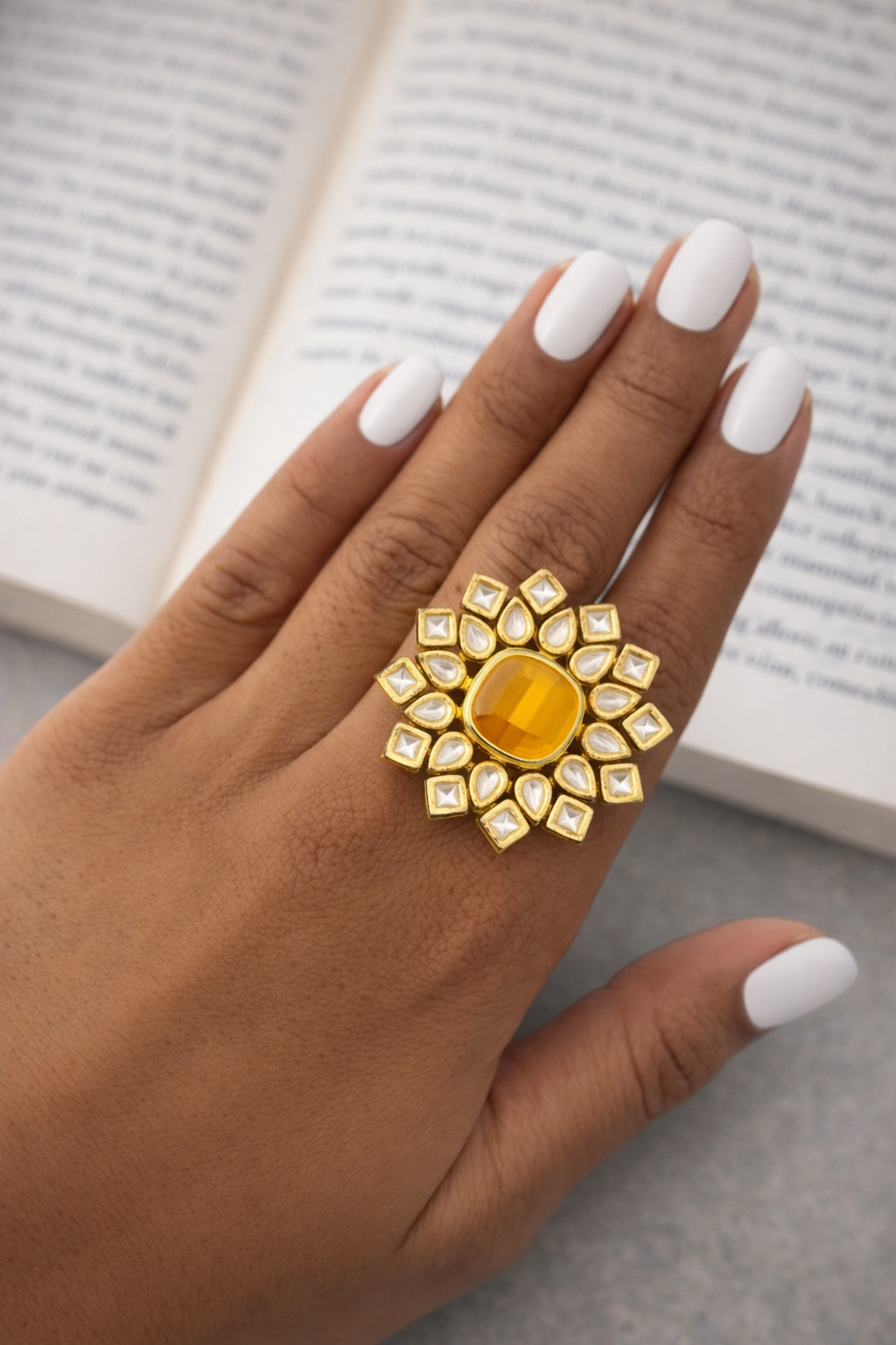 Gold ring with a yellow gemstone on a hand with white nail polish, against a blurred book background.