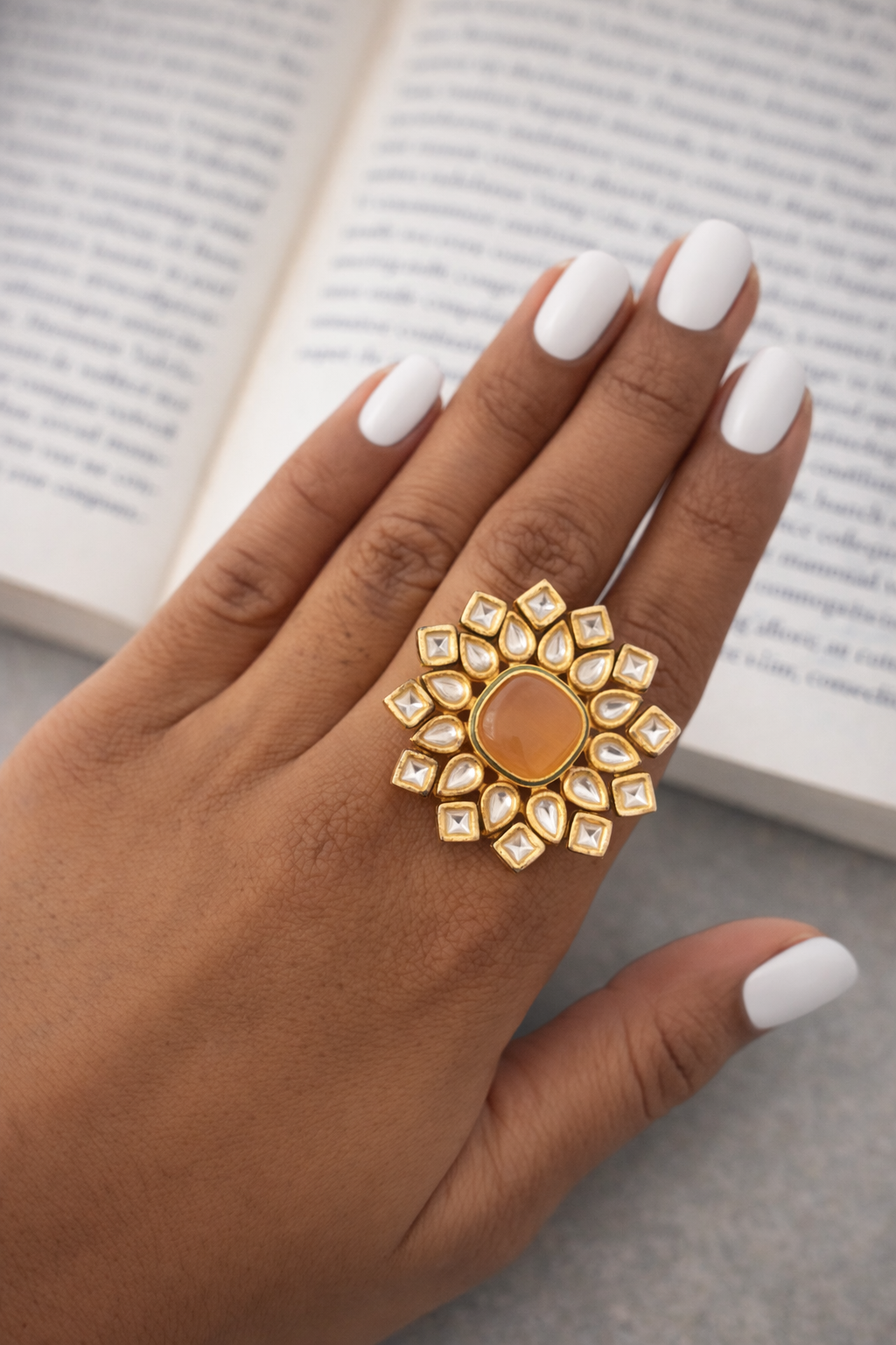 Gold ring with a sunflower design on a hand with white nail polish, against a blurred book background.