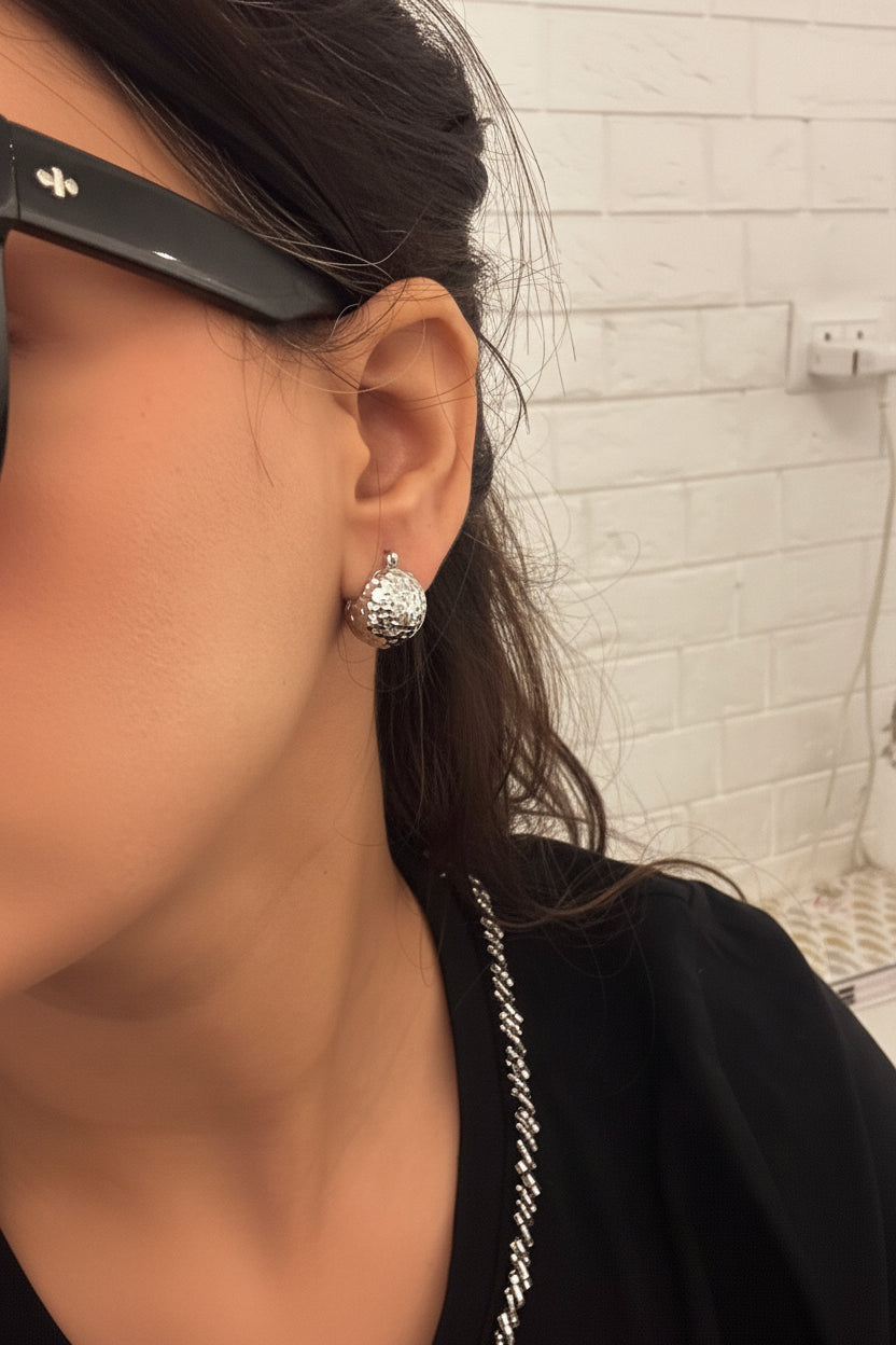 A close-up shot of a person's ear wearing a textured silver stud earring, with dark hair and a black top visible in the background.