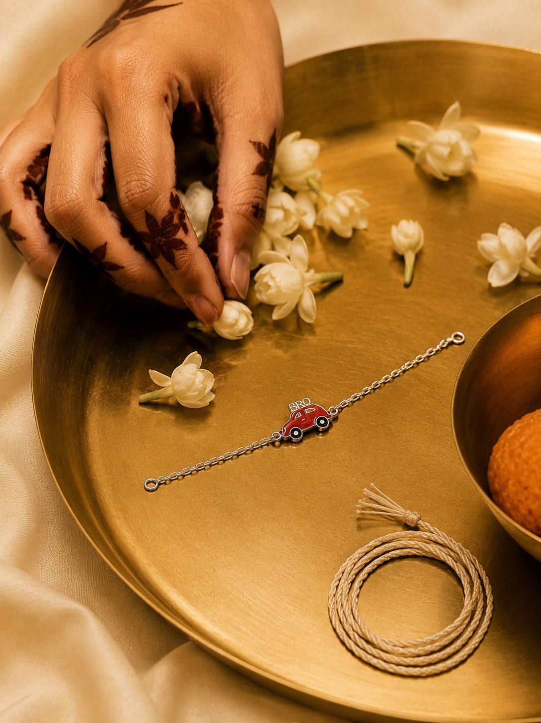 A delicate silver-toned chain bracelet with a small red car charm, displayed on a gold tray alongside white flowers, a coiled cord, and a hand with henna designs.