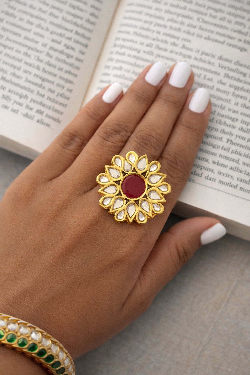 Gold ring with a red gemstone on a hand with white nail polish, against an open book background.