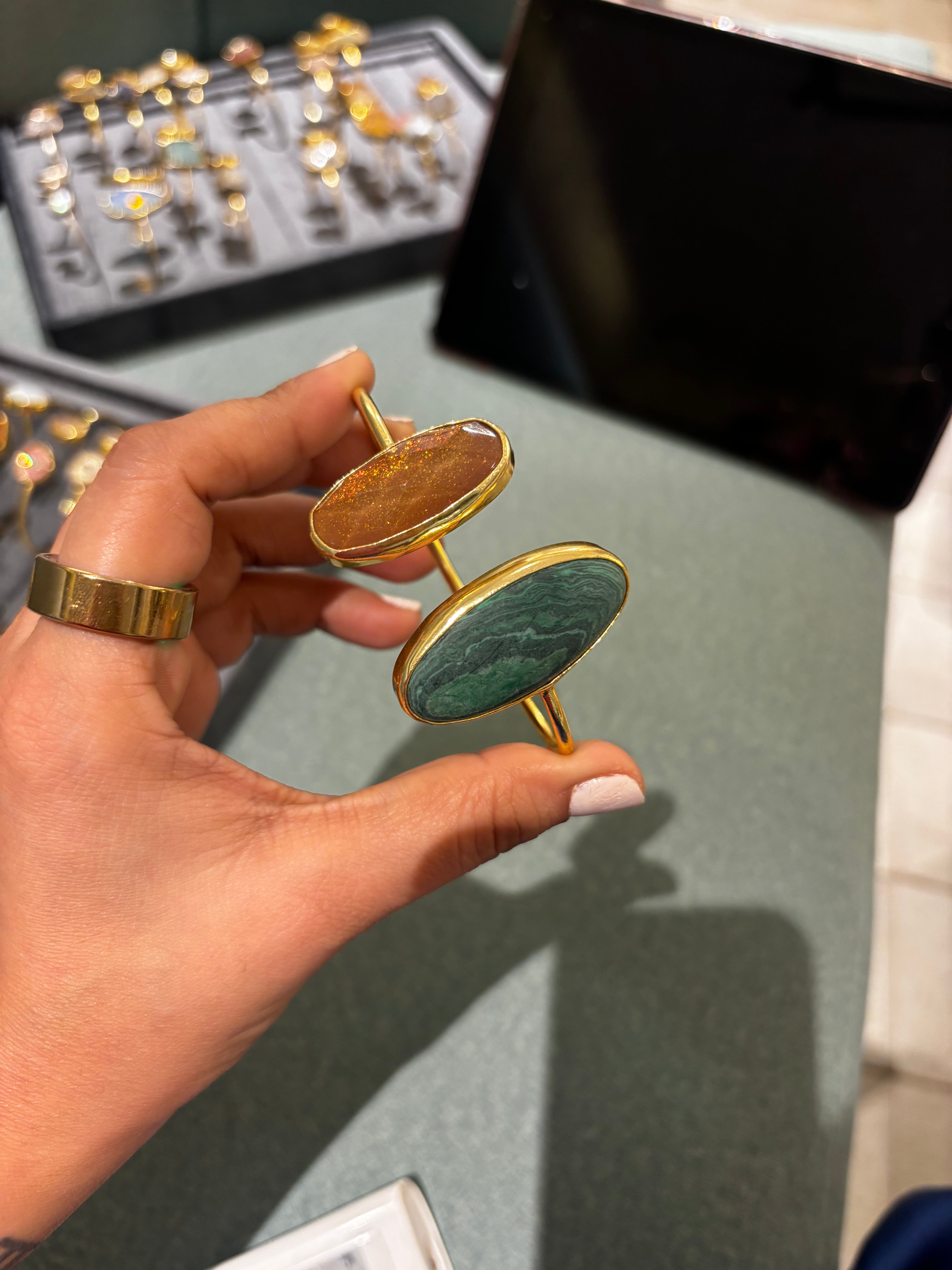 A hand holds a pair of gold-toned oval cufflinks, one with a warm brown stone and the other with a green marbled stone, against a jewelry display background.