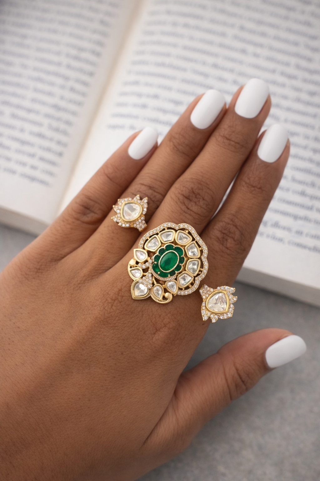 Hand wearing three ornate rings with green gemstones against a blurred book background